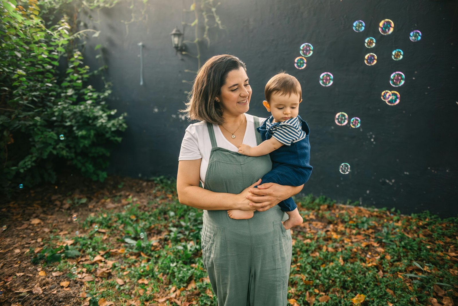 A mother in green overalls holding her baby in a leafy backyard, the two looking at each other as soap bubbles drift past: the calm, everyday warmth Parent Starts is built around.